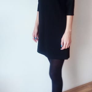 May include: A black mock neck dress with long sleeves and a straight silhouette. The dress is paired with black tights and black heeled boots.