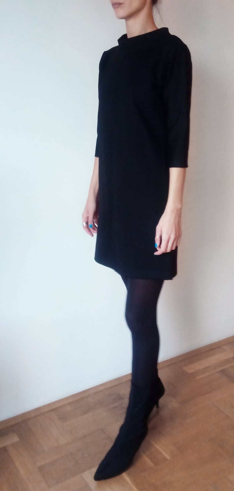 May include: A black mock neck dress with long sleeves and a straight silhouette. The dress is paired with black tights and black heeled boots.