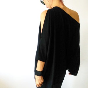 May include: A black, one-shoulder, long-sleeved top with a relaxed fit. The top has a single, long sleeve on the right side and a cut-out shoulder on the left side. The model is wearing black pants and sunglasses.