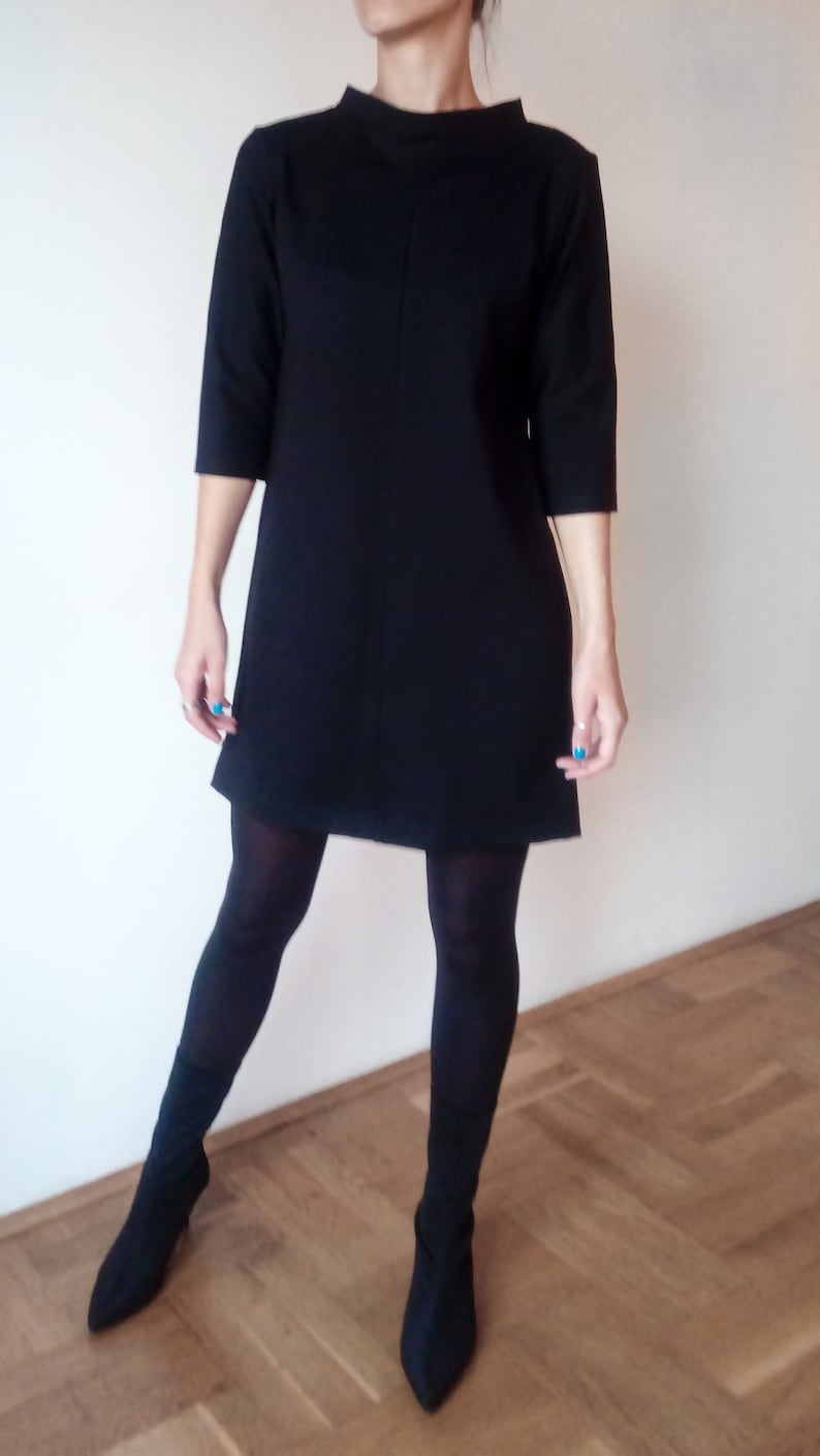 May include: A black shift dress with a mock neck and three-quarter sleeves. The dress is knee-length and has a straight silhouette. The model is wearing black tights and black ankle boots.