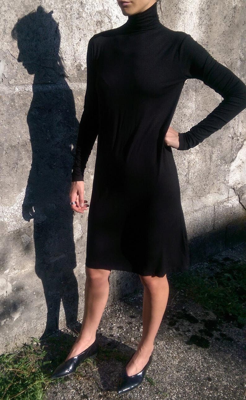 May include: A black turtleneck dress with long sleeves. The dress is fitted and falls to the knee. The model is wearing black pointed-toe shoes.