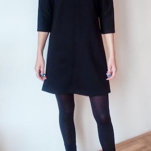 May include: A black shift dress with a mock neck and three-quarter sleeves. The dress is worn with black tights and black heeled ankle boots.