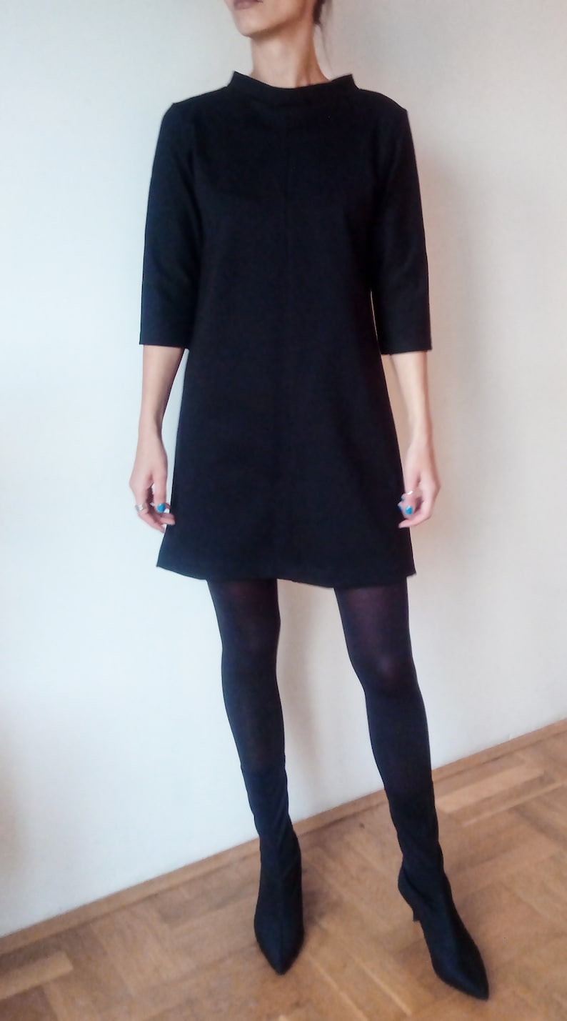 May include: A black shift dress with a mock neck and three-quarter sleeves. The dress is worn with black tights and black heeled ankle boots.