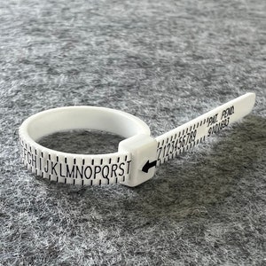 May include: White plastic ring sizer with a ruler and alphabet markings. The ruler goes from 1 to 10 and the alphabet goes from F to T. The ring sizer is on a grey surface.