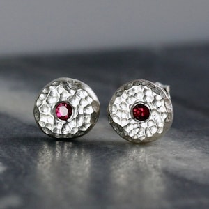May include: A pair of silver stud earrings with a hammered texture and a small red gemstone in the center of each earring.