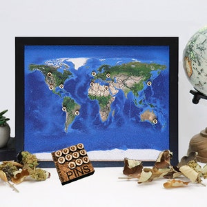 May include: A framed world map with a blue ocean and green landmasses.  There are small gold push pins placed on the map.  A small wooden box with the word "PINS" printed on it is in the lower left corner of the image.  The box contains additional gold push pins.