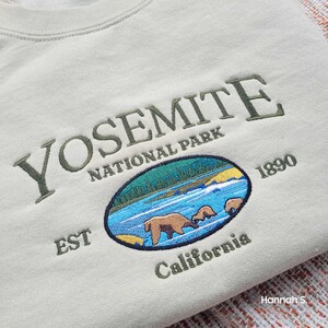 May include: A beige sweatshirt with an embroidered Yosemite National Park logo. The logo features a green, blue and white illustration of a river with three bears, and the text "Yosemite National Park Est 1890 California".