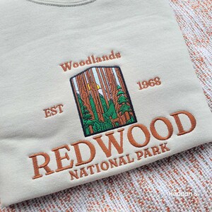 May include: A beige sweatshirt with an embroidered logo for Redwood National Park. The logo features a stylised image of a forest with a sun peeking through the trees, the words "Woodlands" and "EST 1968", and the text "REDWOOD NATIONAL PARK".