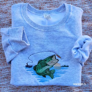 May include: A gray sweatshirt with an embroidered image of a largemouth bass jumping out of the water with a fishing lure in its mouth. The bass is green and has a white belly. The water is blue and there is a sunset in the background.