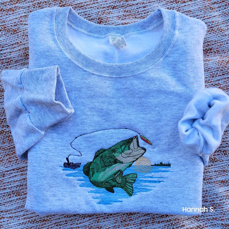 May include: A gray sweatshirt with an embroidered image of a largemouth bass jumping out of the water with a fishing lure in its mouth. The bass is green and has a white belly. The water is blue and there is a sunset in the background.
