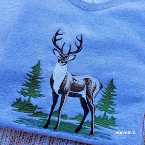 May include: Embroidered gray sweatshirt with a brown and white deer standing in a green forest scene. The text "Hannah S." is in the bottom right corner.