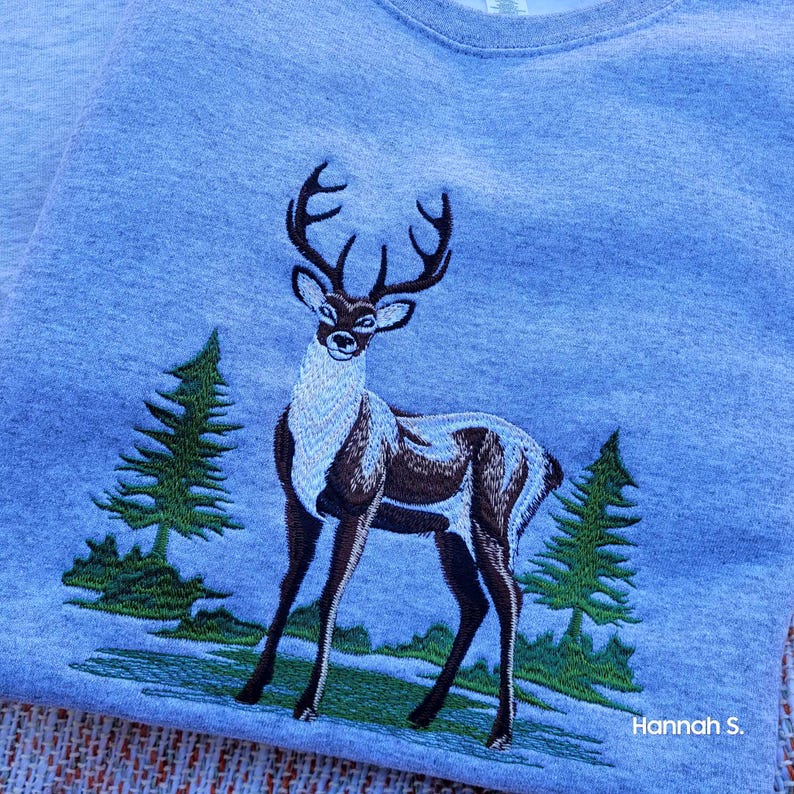 May include: Embroidered gray sweatshirt with a brown and white deer standing in a green forest scene. The text "Hannah S." is in the bottom right corner.