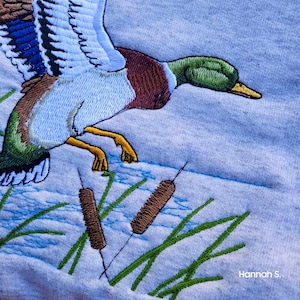 May include: Embroidered design of a mallard duck flying over a pond with cattails. The duck is blue, green, brown, and white. The cattails are brown and green. The background is a light blue.