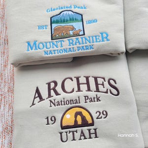 May include: Two beige sweatshirts with embroidered national park logos. The top sweatshirt features a blue and white logo for Mount Rainier National Park with a brown bear and cub. The bottom sweatshirt features a brown logo for Arches National Park with a brown and yellow arch and the text "UTAH".