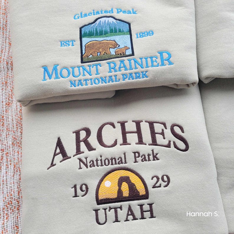 May include: Two beige sweatshirts with embroidered national park logos. The top sweatshirt features a blue and white logo for Mount Rainier National Park with a brown bear and cub. The bottom sweatshirt features a brown logo for Arches National Park with a brown and yellow arch and the text "UTAH".