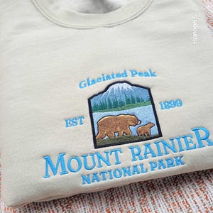 May include: A light beige sweatshirt with a blue embroidered design featuring a mountain, two bears, and the text "Glaciated Peak EST 1899 MOUNT RAINIER NATIONAL PARK".