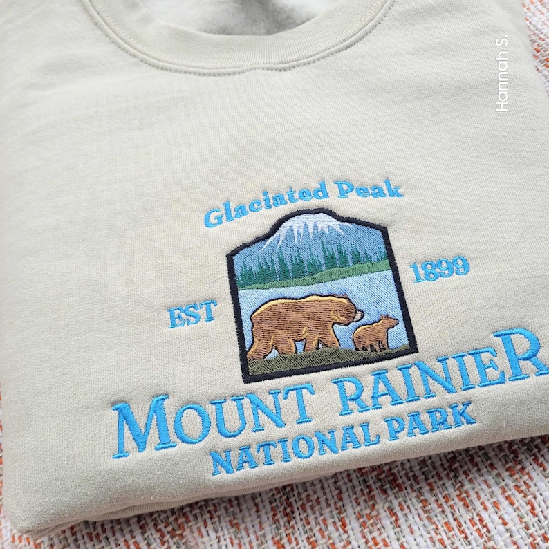 May include: A light beige sweatshirt with a blue embroidered design featuring a mountain, two bears, and the text "Glaciated Peak EST 1899 MOUNT RAINIER NATIONAL PARK".