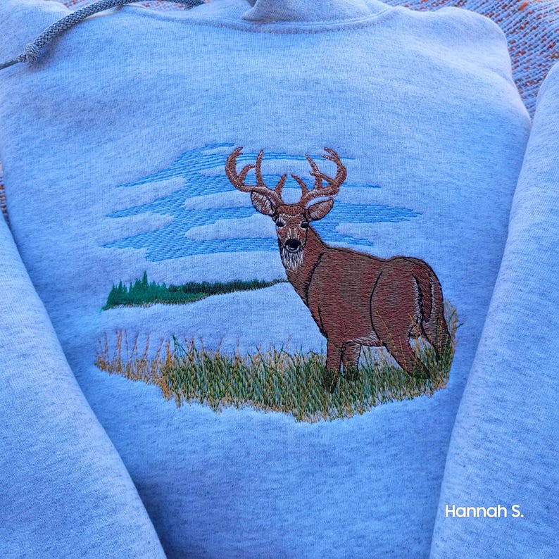 May include: A gray hooded sweatshirt with an embroidered image of a brown deer with antlers standing in a field of green grass. The deer is looking to the left. The background is a blue sky with white clouds.