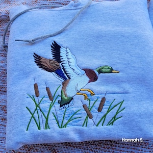 May include: A gray hooded sweatshirt with an embroidered image of a mallard duck in flight over a pond with cattails. The duck is brown, green, and blue. The text "Hannah S." is embroidered in the bottom right corner.