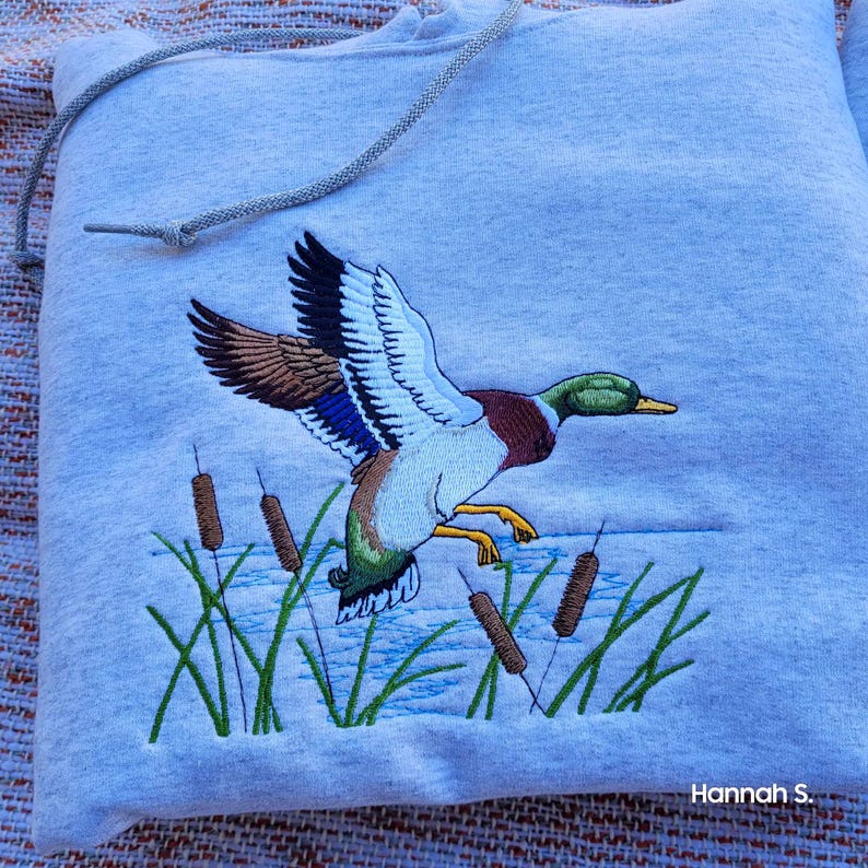 May include: A gray hooded sweatshirt with an embroidered image of a mallard duck in flight over a pond with cattails. The duck is brown, green, and blue. The text "Hannah S." is embroidered in the bottom right corner.