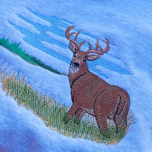 May include: Embroidered design of a brown deer with large antlers standing in a field of green grass against a blue sky background.