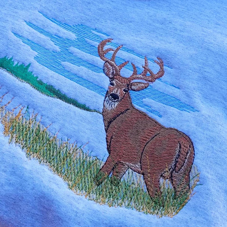May include: Embroidered design of a brown deer with large antlers standing in a field of green grass against a blue sky background.