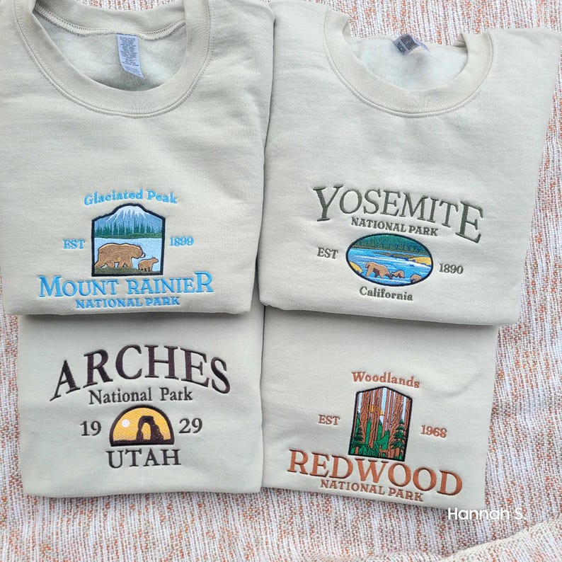 May include: Four beige sweatshirts with embroidered national park logos. The top left sweatshirt features Mount Rainier National Park, the top right sweatshirt features Yosemite National Park, the bottom left sweatshirt features Arches National Park, and the bottom right sweatshirt features Redwood National Park.