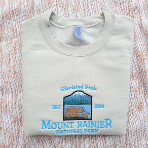 May include: A beige sweatshirt with an embroidered logo for Mount Rainier National Park. The logo features a mountain scene with two bears, the text "Glaciated Peak", "EST 1899", and "Mount Rainier National Park".