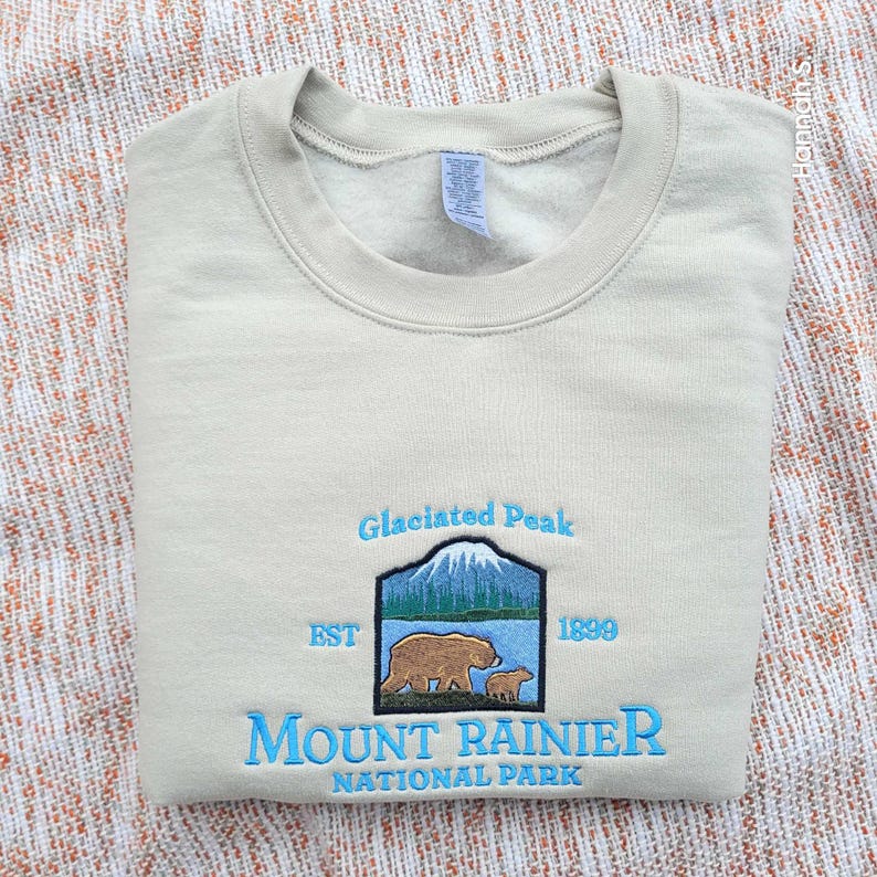 May include: A beige sweatshirt with an embroidered logo for Mount Rainier National Park. The logo features a mountain scene with two bears, the text "Glaciated Peak", "EST 1899", and "Mount Rainier National Park".