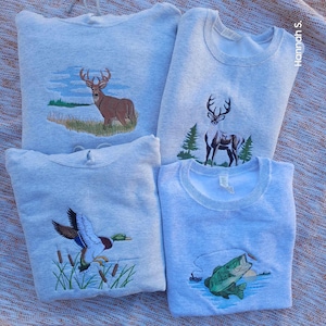 May include: Four gray sweatshirts with embroidered designs. One sweatshirt features a deer, another a duck, another a fish, and the last a deer. The sweatshirts are folded and stacked on top of each other.
