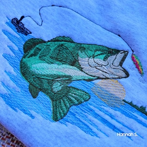 May include: Embroidered design of a green bass fish jumping out of the water with a lure in its mouth. The fish is on a blue background with a yellow sun and blue water ripples.