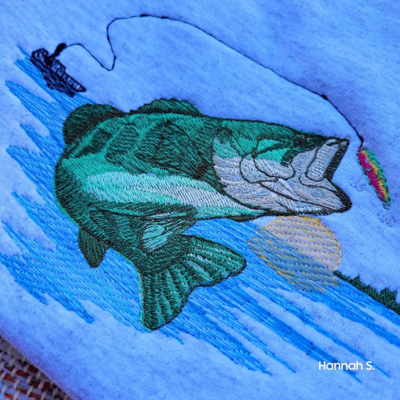May include: Embroidered design of a green bass fish jumping out of the water with a lure in its mouth. The fish is on a blue background with a yellow sun and blue water ripples.