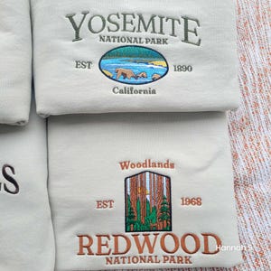 May include: Two beige sweatshirts with embroidered national park logos. The top sweatshirt features the Yosemite National Park logo with a bear and cubs, established in 1890. The bottom sweatshirt features the Redwood National Park logo with a forest scene, established in 1968.