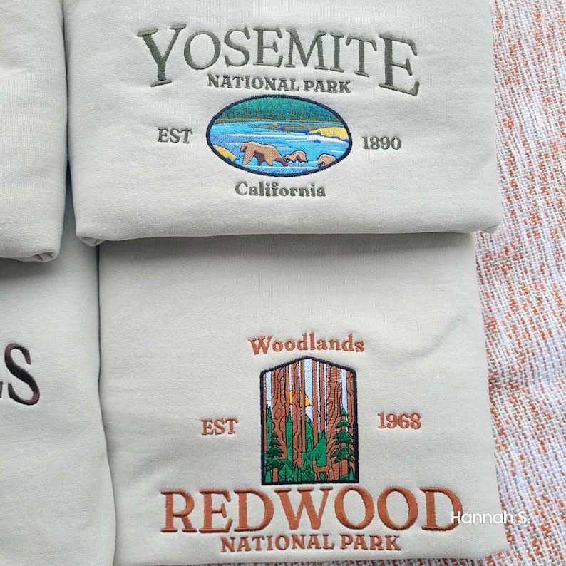 May include: Two beige sweatshirts with embroidered national park logos. The top sweatshirt features the Yosemite National Park logo with a bear and cubs, established in 1890. The bottom sweatshirt features the Redwood National Park logo with a forest scene, established in 1968.