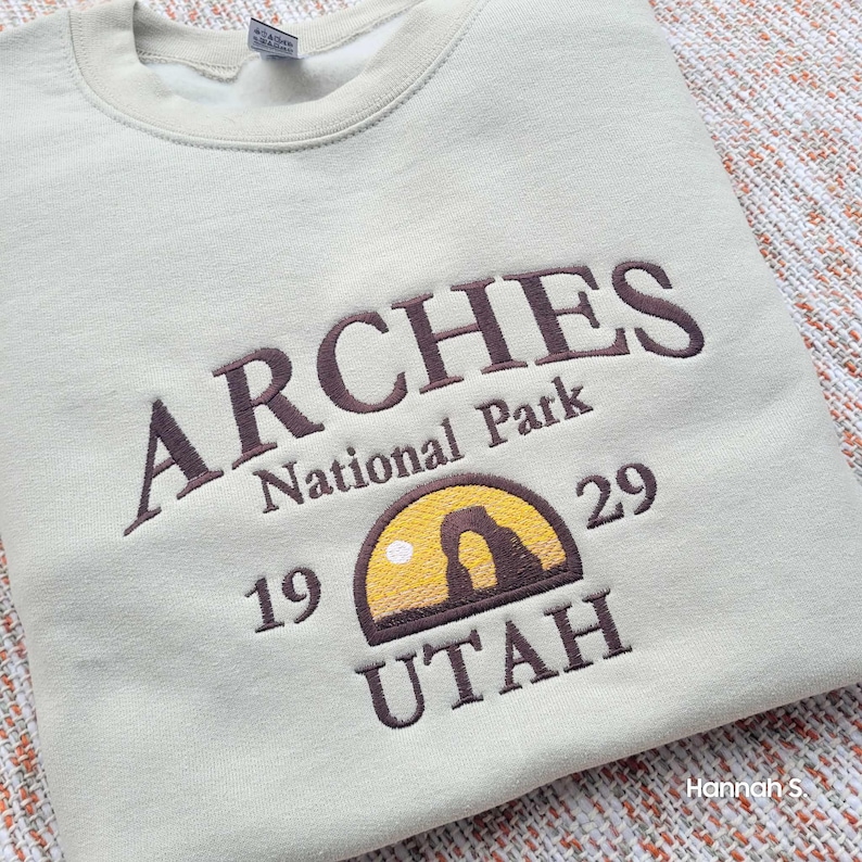 May include: A light beige sweatshirt with brown embroidered text that reads "ARCHES National Park 19 29 UTAH". The text is arched and the logo features a stylised depiction of a sunset with a rock formation.