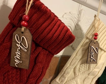 May include: Four Christmas stockings with personalized wooden tags. The stockings are red, white, green, and red. The tags are brown with white lettering. The names on the tags are "Shawn", "Zach", "Max", and "Lily".