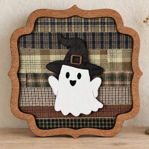 May include: A decorative wall sign featuring a white ghost wearing a black witch hat with a copper-colored buckle. The ghost is centered on a background of horizontal fabric strips in various plaid patterns of brown, tan, and navy blue, all encased in a scalloped wooden frame.