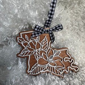 May include: A wooden ornament shaped like the state of Louisiana with a white magnolia flower design. The ornament is attached to a black and white checkered ribbon.
