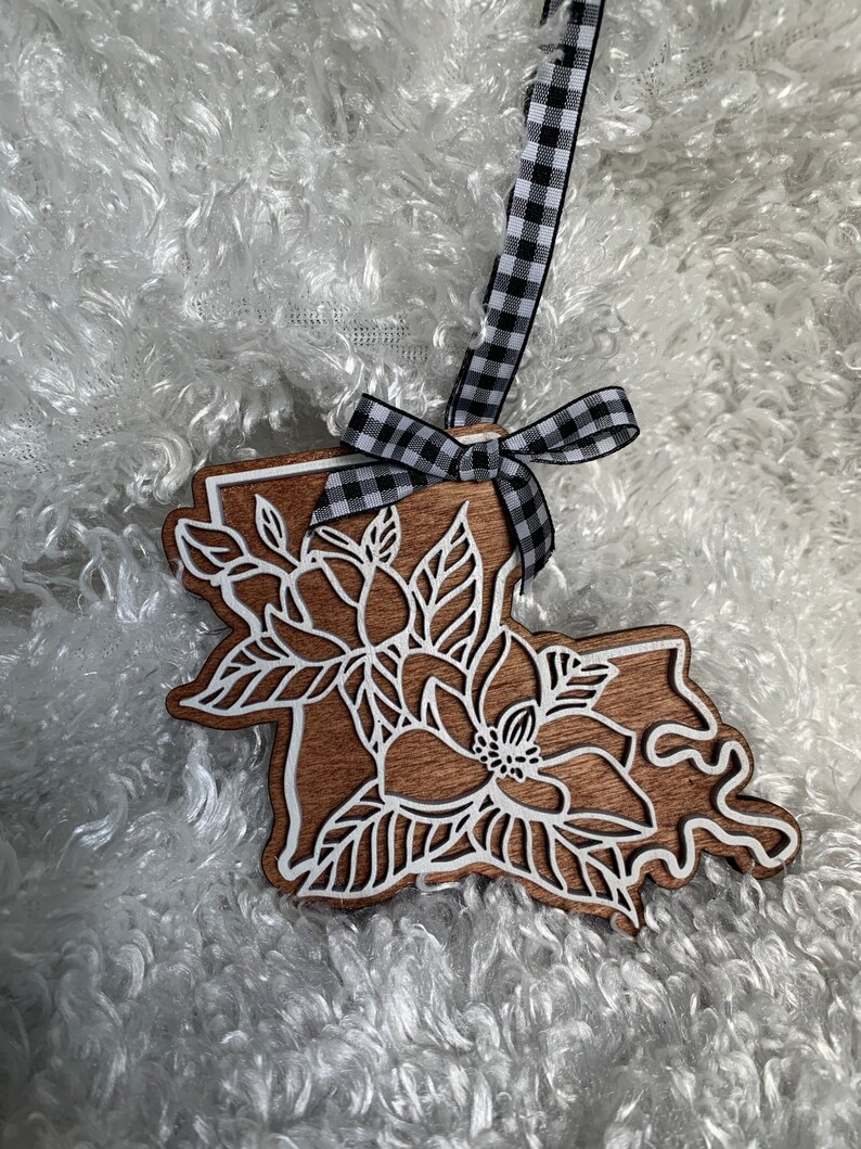 May include: A wooden ornament shaped like the state of Louisiana with a white magnolia flower design. The ornament is attached to a black and white checkered ribbon.