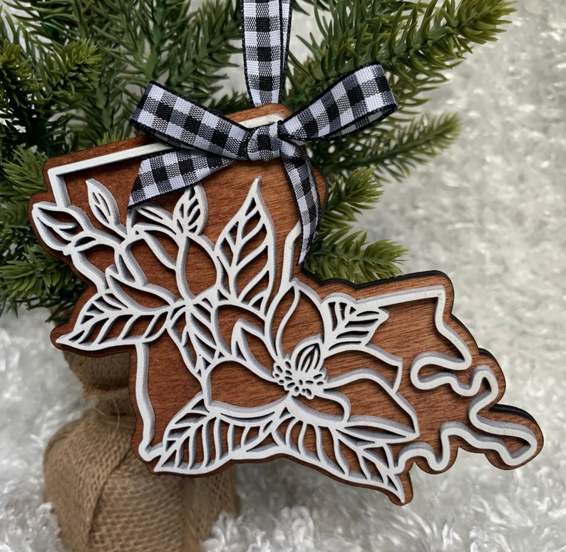 May include: A wooden ornament shaped like the state of Louisiana with a white magnolia flower design. The ornament is decorated with a black and white checkered ribbon.