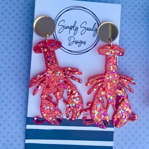 May include: A pair of coral-colored lobster earrings with glitter accents. The earrings are made of a translucent material and feature a gold-colored circular stud. The earrings are displayed on a white card with the text "Simply Sandy Designs".