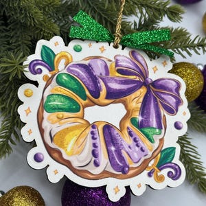 May include: A wooden ornament shaped like a King Cake, featuring purple, green, and yellow icing. The ornament has a green glitter bow and a gold string for hanging. Decorative elements include fleur-de-lis and small dots.