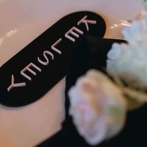 May include: A black place card with the name "KELSEY" in white, placed on a white plate. The card is rectangular with rounded edges. Soft focus flowers and a black napkin are in the background.