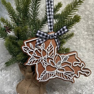 May include: Wooden Christmas ornament shaped like the state of Louisiana with a white magnolia flower design. The ornament is hanging from a black and white checkered ribbon.
