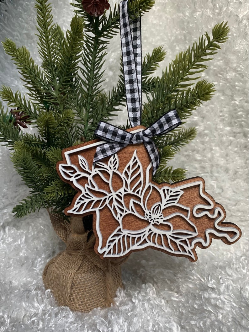 May include: Wooden Christmas ornament shaped like the state of Louisiana with a white magnolia flower design. The ornament is hanging from a black and white checkered ribbon.