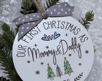 May include: A round white Christmas ornament featuring the black text "OUR FIRST CHRISTMAS AS Mommy & Daddy" and "2026". The design includes three small green glittery Christmas trees, three small hearts, and snowflakes. A grey polka dot ribbon is tied at the top.