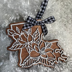 May include: A wooden ornament shaped like the state of Louisiana with a white magnolia flower design. The ornament is decorated with a black and white checkered ribbon.