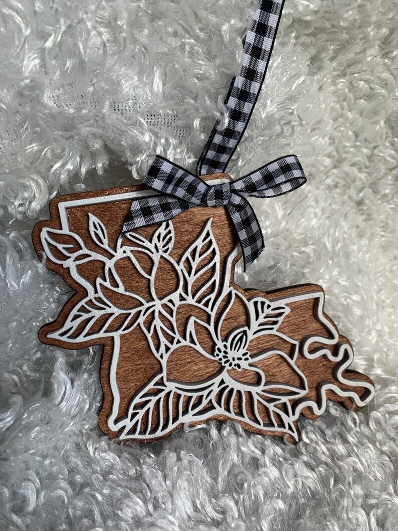 May include: A wooden ornament shaped like the state of Louisiana with a white magnolia flower design. The ornament is decorated with a black and white checkered ribbon.