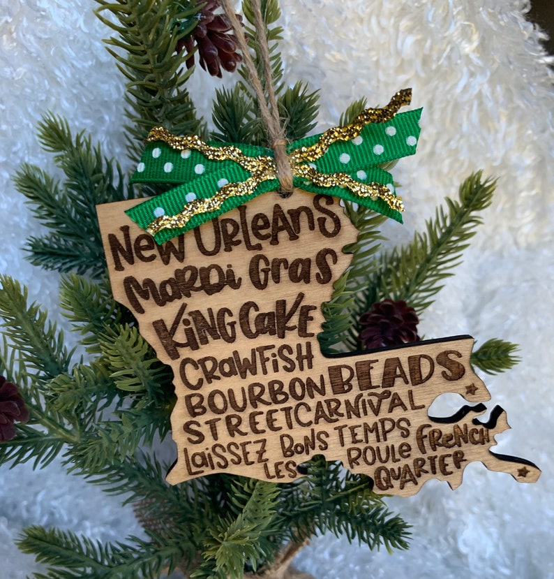 May include: A wooden ornament shaped like the state of Louisiana with the words "New Orleans Mardi Gras" and other related phrases written on it. The ornament is decorated with a green and gold ribbon.