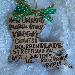 May include: Wooden ornament shaped like the state of Louisiana with the words "New Orleans Mardi Gras King Cake Crawfish Bourbon Beads Streetcarnival Laissez Les Bons Temps Roule French Quarter" written on it. The ornament is decorated with a green and gold ribbon.